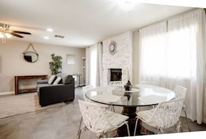 Ein Wohnzimmer mit rundem Glastisch, weißen Stühlen, einem grauen Sofa, einem Kamin und einem Deckenventilator.