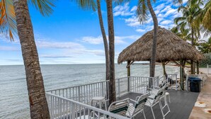 Eine Strandterrasse mit weißen Stühlen, einer Struktur mit Reetdach und Palmen.