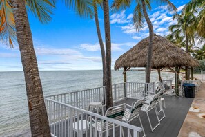 Eine Strandterrasse mit weißen Stühlen, einer Struktur mit Reetdach und Palmen.
