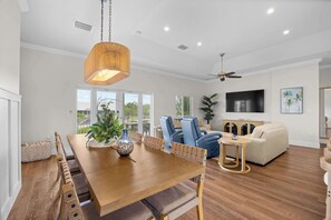 Un vaste espace de vie comprenant une table à manger en bois, des chaises bleues, un canapé blanc et un téléviseur fixé au mur.
