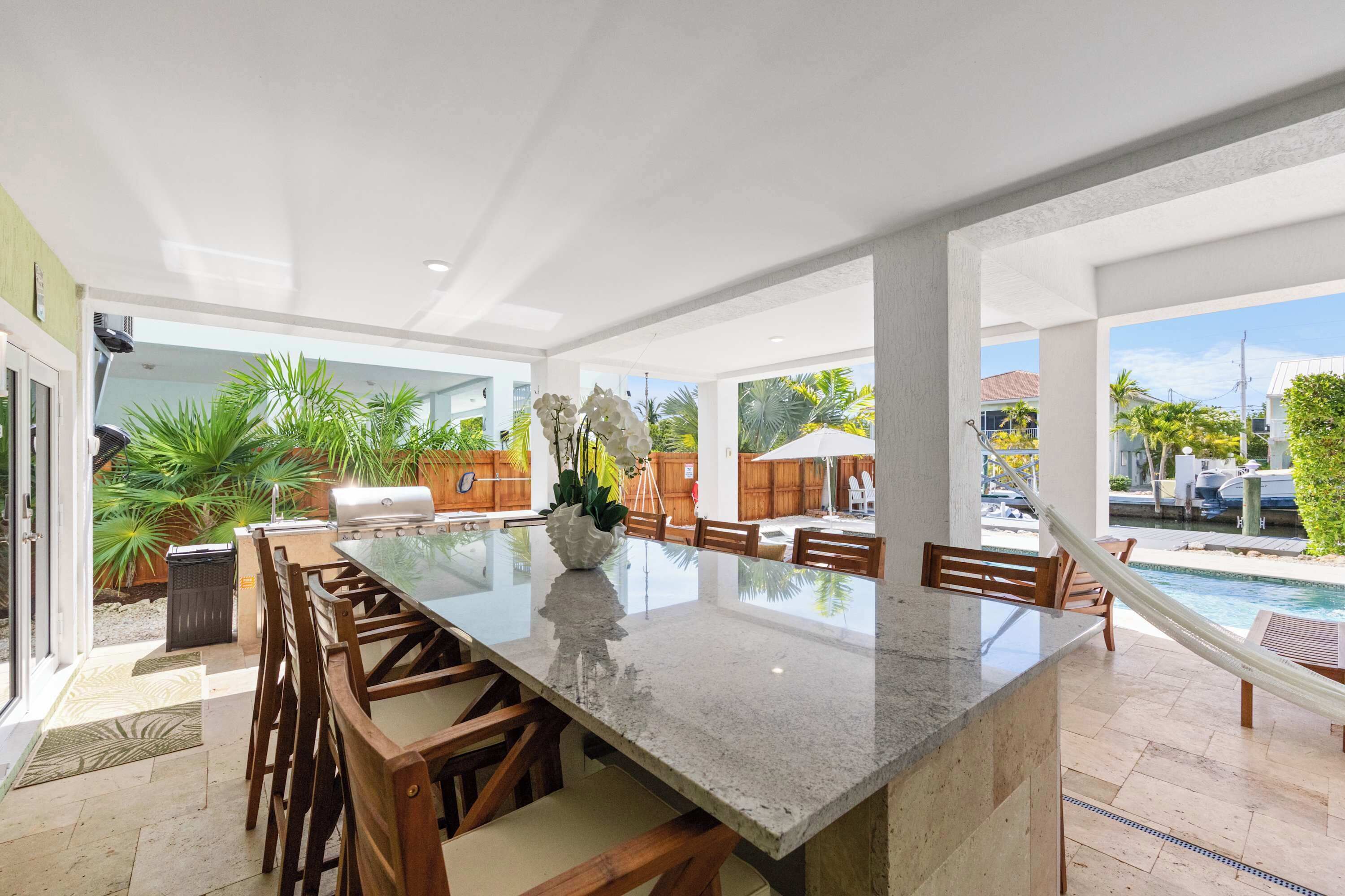 A spacious outdoor dining area with a large granite table, wooden chairs, and a view of a swimming pool and greenery.