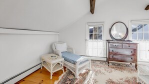 A room with a white wicker chair, a bench with a striped cushion, a wooden dresser with a mirror, and a patterned rug.
