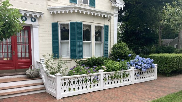 A white building with green shutters, a red door, and a white picket fence with plants.