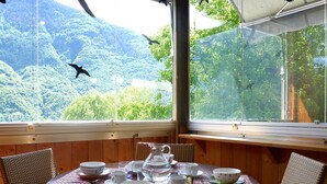 Une pièce aux murs lambrissés, avec une table préparée pour le thé, un fauteuil en osier et une vue sur une colline boisée.