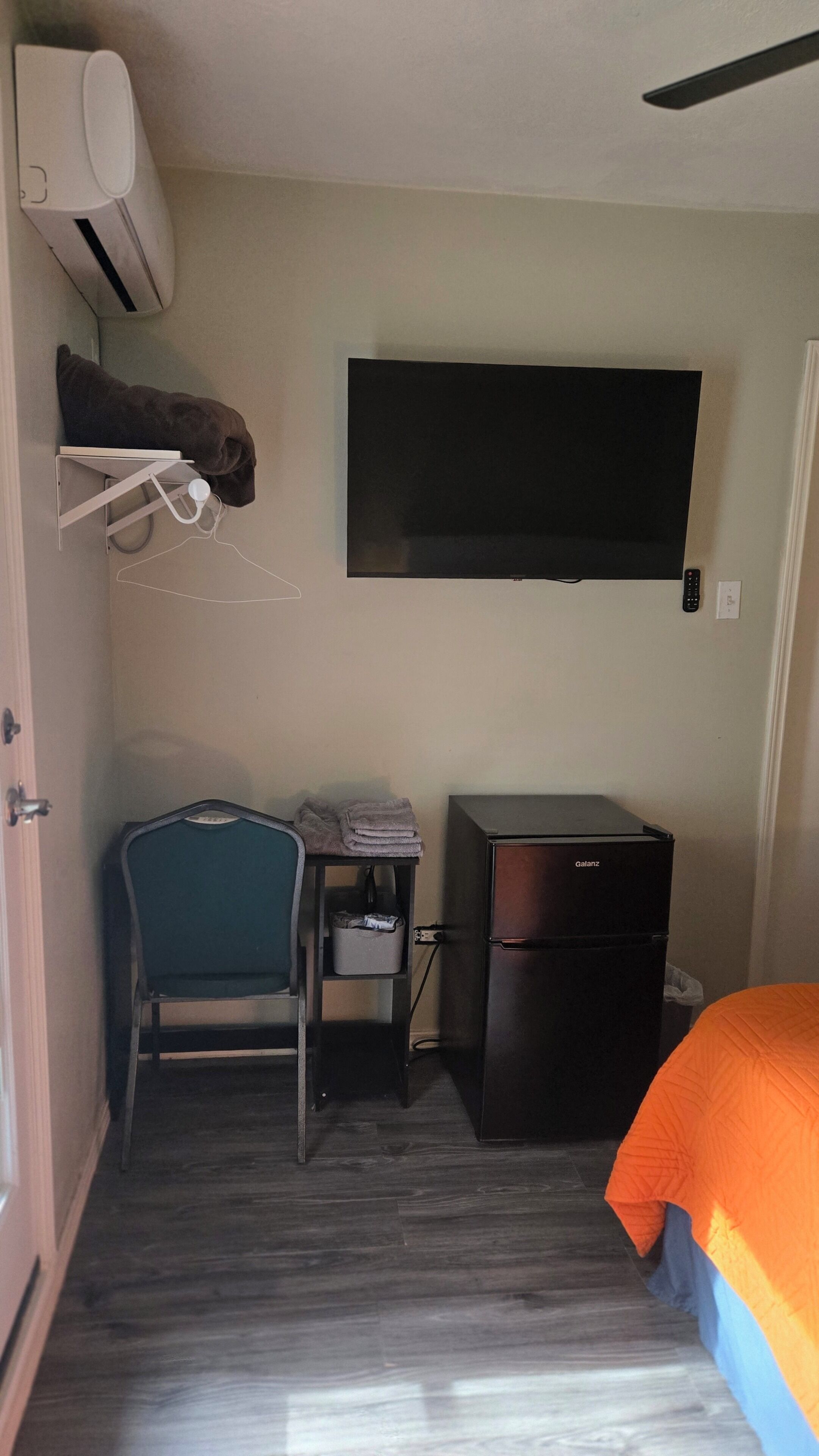 Ein kleines Zimmer mit Schreibtisch, Stuhl, Bett und einem an der Wand montierten Flachbildfernseher.
