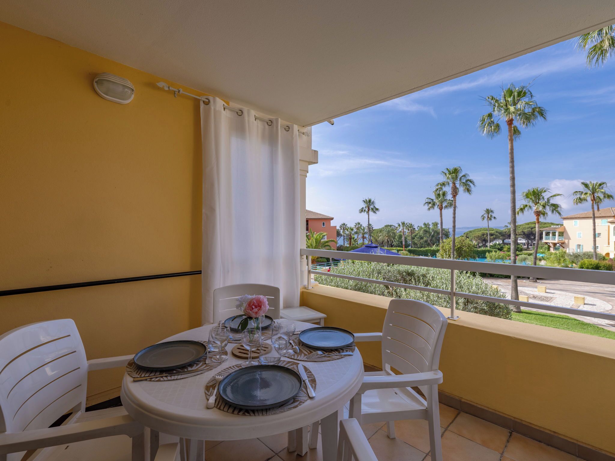 A balcony with a table set for dining, white chairs, a white curtain, and a view of palm trees and buildings.
