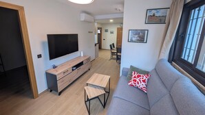 Ein modernes Wohnzimmer mit Flachbildfernseher, einer Holz-Unterhaltungseinheit, einem grauen Sofa und einem Essbereich im Hintergrund.