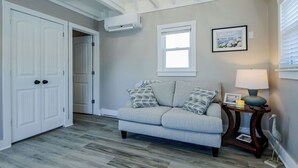 A room with a grey couch, a small table with a lamp, a framed picture on the wall, and a white door.