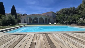 Ein Schwimmbecken mit Holzterrasse, zwei Liegestühlen und einem Gebäude mit Bögen im Hintergrund.