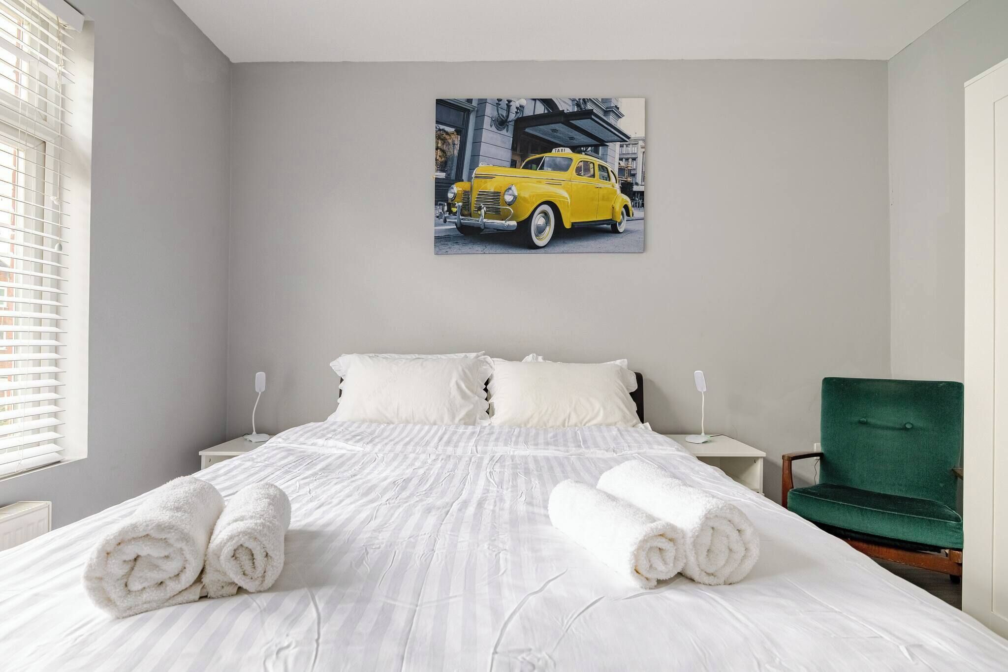 Une chambre d’hôtel avec un lit, du linge de lit blanc, un fauteuil vert et un tableau représentant une voiture jaune accroché au mur.