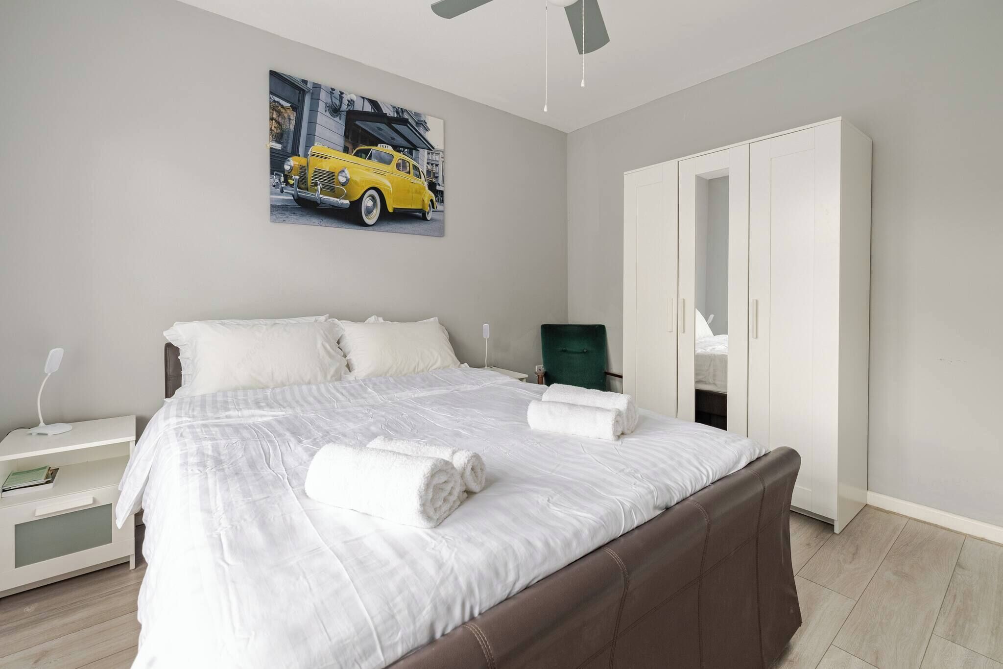 Ein Schlafzimmer mit einem Bett, weißer Bettwäsche, einem Nachttisch, einem Schreibtisch, einem Stuhl, einem Kleiderschrank und einem Deckenventilator.