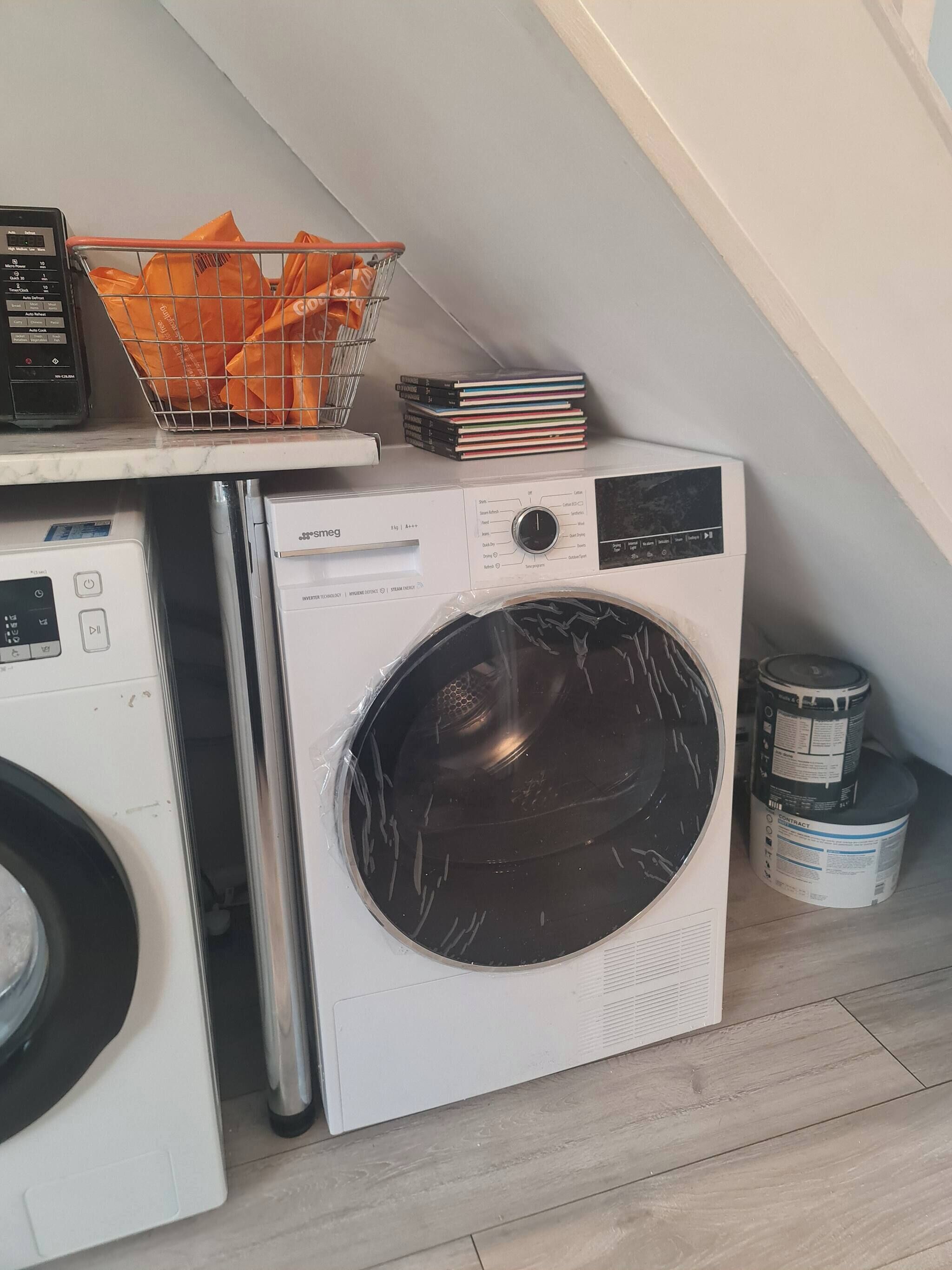 A compact laundry area with a front-loading washer and dryer, a metal basket with orange items, and stacked books.
