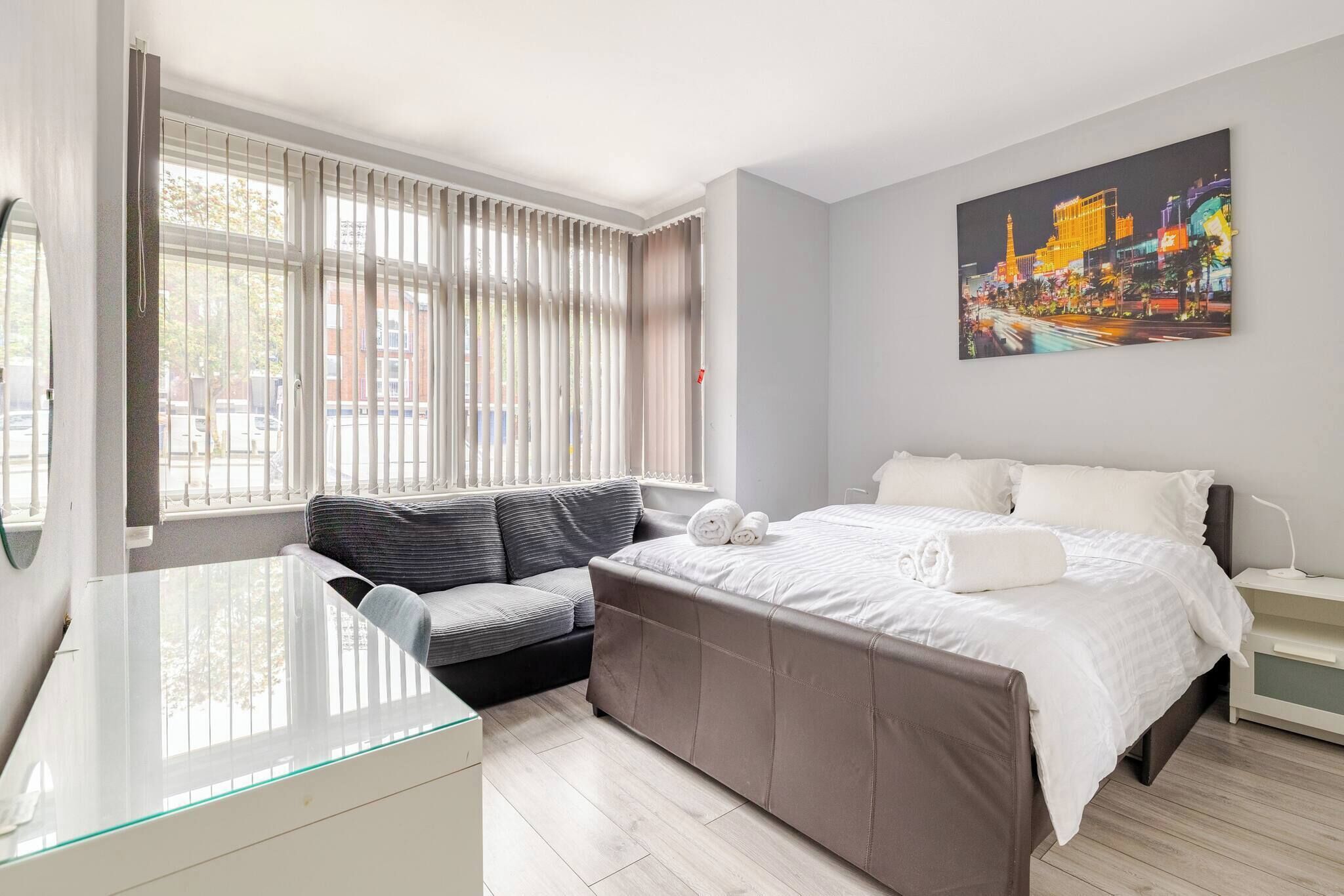 A modern bedroom with a bed, a sofa, a desk, and a cityscape painting.