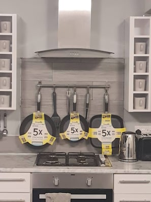 A kitchen with a gas stove, three frying pans hanging on a rack, and a kettle on the counter.