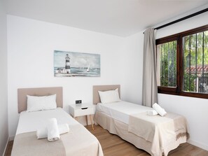 A hotel room with two beds, a nightstand, a painting of a lighthouse, and a window with a view of greenery. - Ca Maestral by Interhome (Marbella)