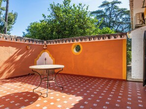 Um pátio com uma mesa redonda e cadeiras, uma parede decorativa com um emblema circular e vista para árvores e um edifício ao fundo.