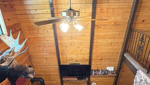 Ein rustikaler Innenraum einer Blockhütte mit Deckenventilator, Ledersofas, Kamin und einem aufgehängten Elchgehörn.