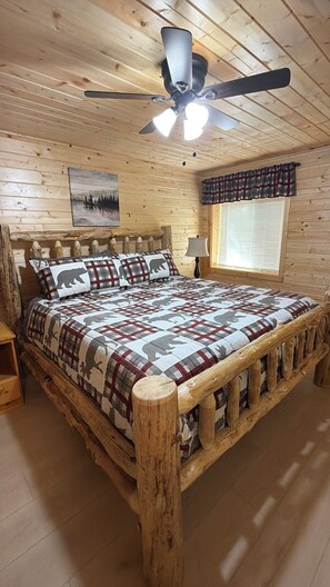 A wooden cabin bedroom with a bed, a ceiling fan, a window with blinds, and a painting on the wall. - 20 min from Yellowstone NP, hot tub, sleeps 18, 3-bedroom, 2.5 bath, Island Park (Island Park)
