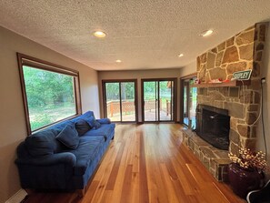 A spacious living room with a blue sofa, a stone fireplace, large windows, and a view of a deck.