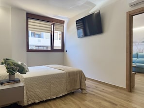 Een slaapkamer met een bed, een aan de muur bevestigde televisie, een raam met zonwering en uitzicht op een andere kamer.