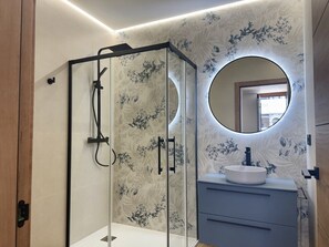 Un bagno moderno con cabina doccia in vetro, uno specchio rotondo e un lavandino bianco.