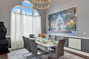 A dining area with a table set for six, a chandelier, a large window with curtains, and a wall-mounted painting of a fountain. - Savannah Family Hub: Game RM, Screened Porch & Work From Home, 15 mins Downtown (Pooler)