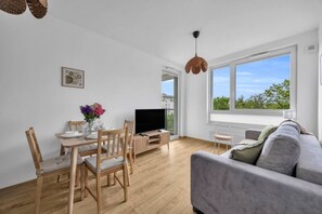 Ein modernes Wohnzimmer mit grauem Sofa, Esstisch aus Holz und Stühlen, einem Flachbildfernseher und großen Fenstern mit Blick auf Grünflächen.