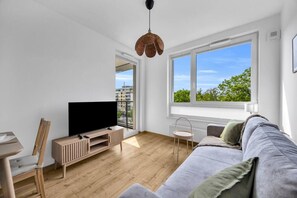 Un salon moderne doté d’un téléviseur à écran plat, de meubles en bois, d’un canapé gris et d’une grande fenêtre offrant une vue sur les bâtiments et les arbres.