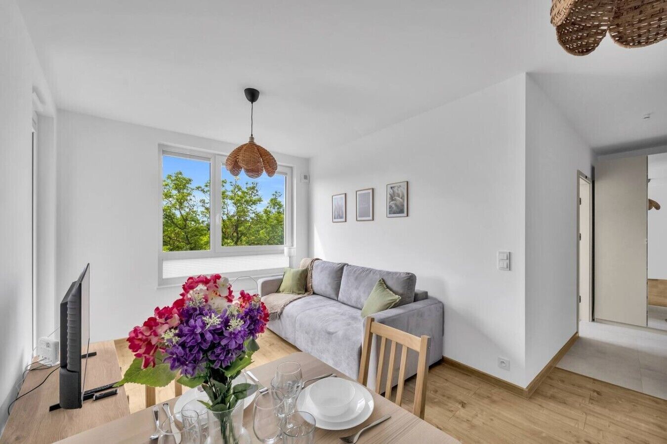 Ein modernes Wohnzimmer mit einem grauen Sofa, einem Esstisch gedeckt mit Tellern und Gläsern, einem Fernseher und einem großen Fenster mit Blick auf Bäume.