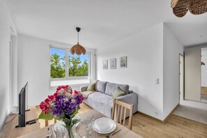 Ein modernes Wohnzimmer mit einem grauen Sofa, einem Esstisch gedeckt mit Tellern und Gläsern, einem Fernseher und einem großen Fenster mit Blick auf Bäume.