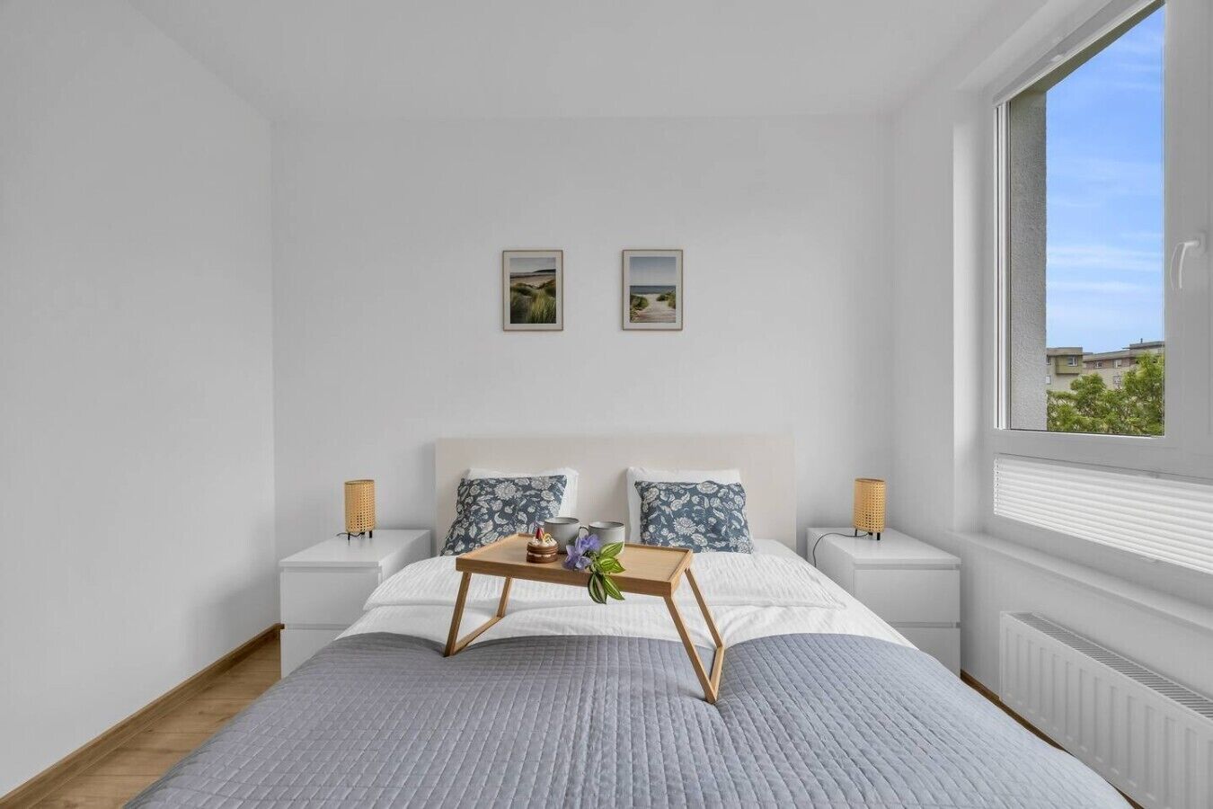 A modern bedroom with a large bed, two bedside tables, a window with a view, and two framed pictures on the wall.
