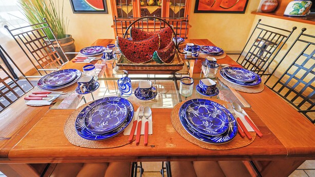 Una mesa de comedor dispuesta con platos azules, servilletas rojas y cubiertos, rodeada de sillas.