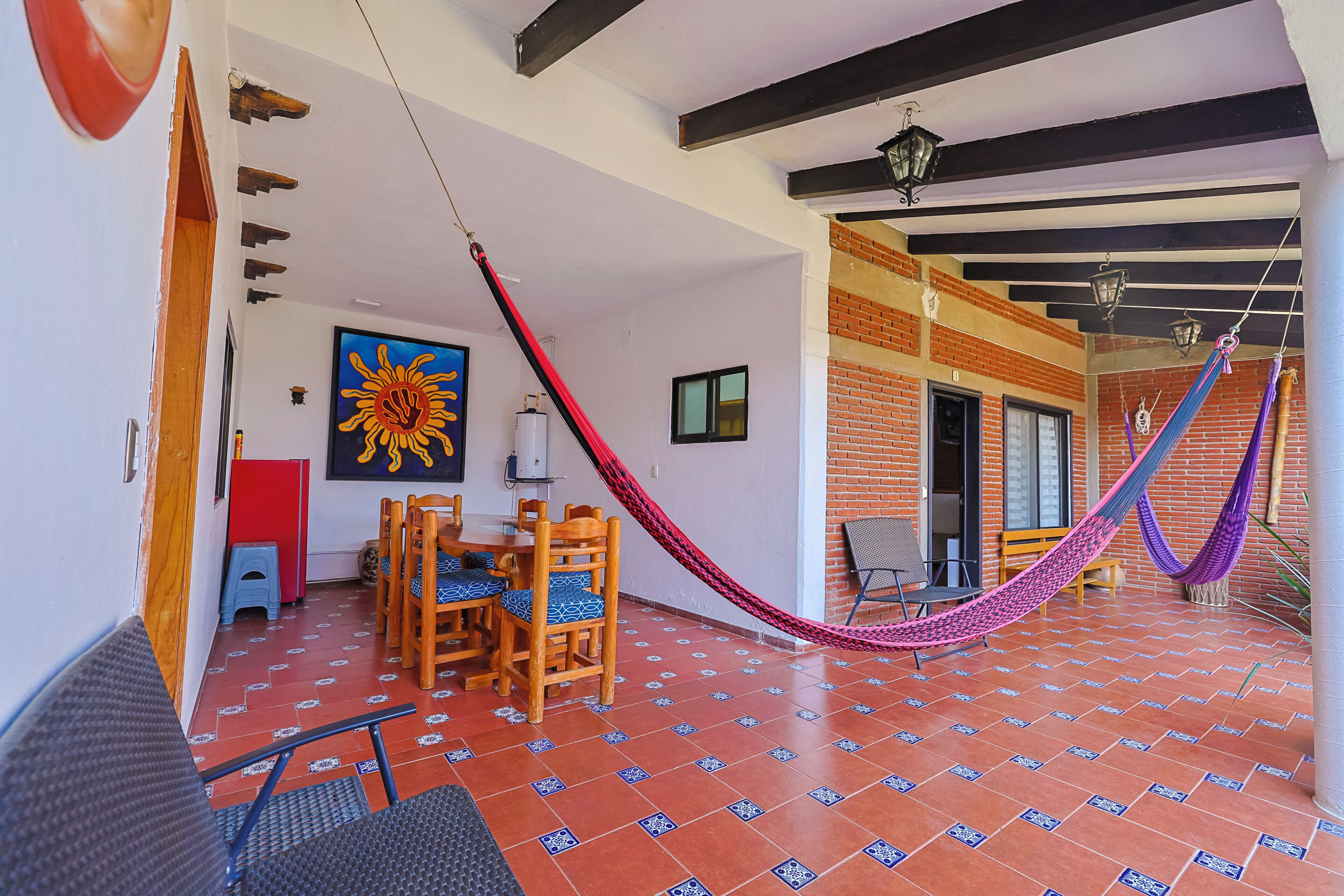 A room with a tiled floor, a wooden dining table with chairs, a hammock, and a large sun painting.