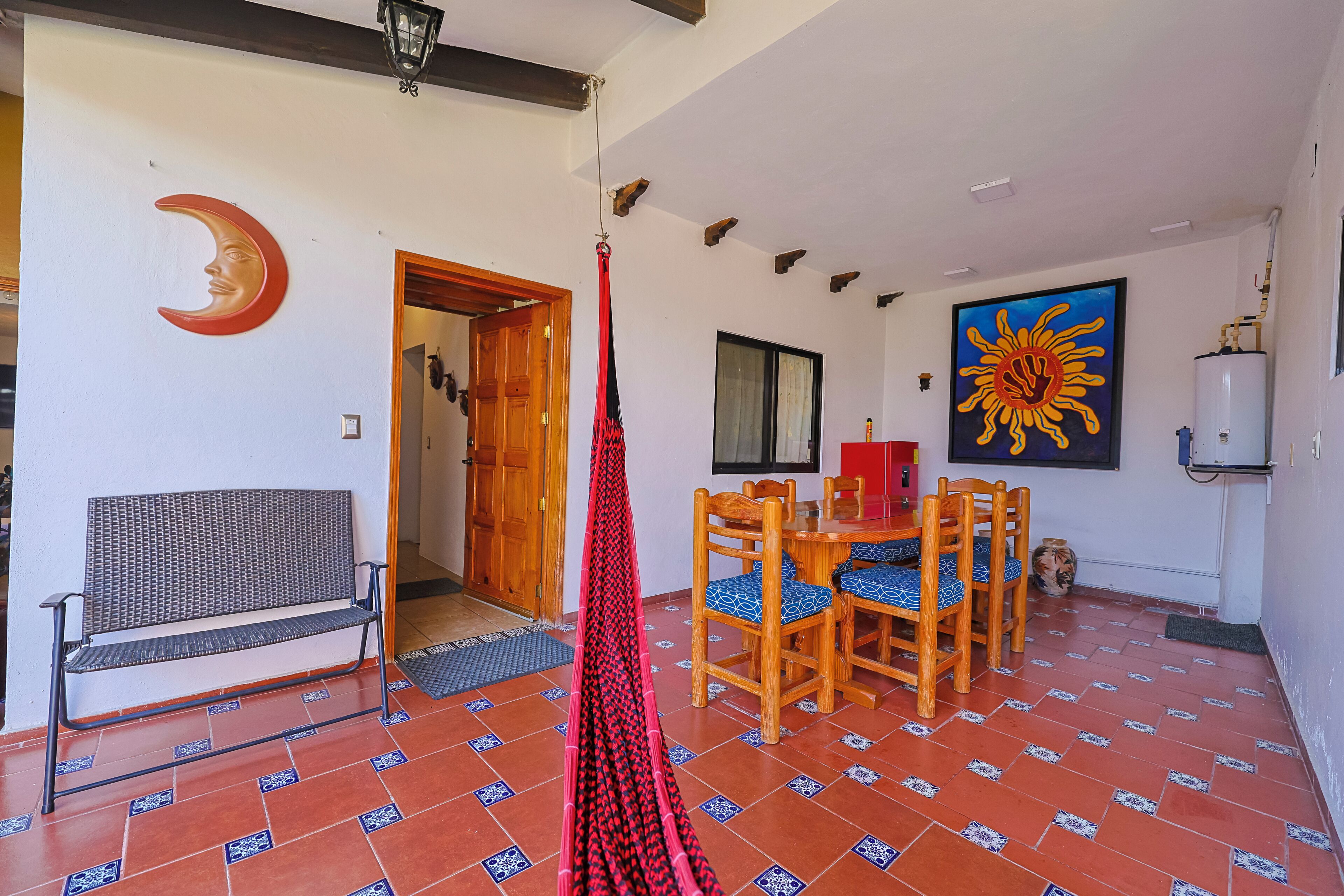 A room with a bench, a dining table with chairs, a hanging hammock, and a large sun painting on the wall.