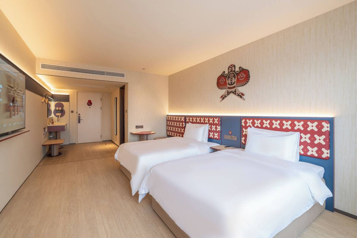 Ein Hotelzimmer mit zwei Betten, einem kleinen Tisch, einer Bank, einem Spiegel und einem an der Wand hängenden Kunstwerk.