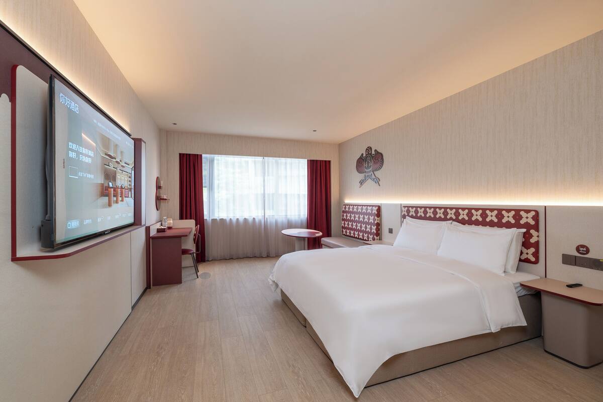 Ein Hotelzimmer mit einem großen Bett, einem Schreibtisch, einem Sessel, einem an der Wand befestigten Fernseher und einem Fenster mit Vorhängen.