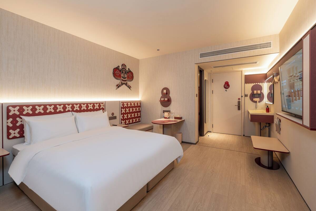 Ein Hotelzimmer mit einem großen Bett, einem Nachttisch, einem kleinen Schreibtisch, einem Spiegel und einer Tür zum Badezimmer.