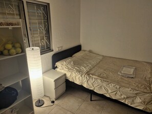 A single bed with a quilt, a bedside table, a lamp, and a window with metal bars. - The Lemon House (Tel Aviv)