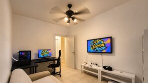 Et værelse med gaming-setup bestående af to skærme, et skrivebord, en stol, en sofa, en loftventilator, et fjernsyn og et hvidt konsolbord.