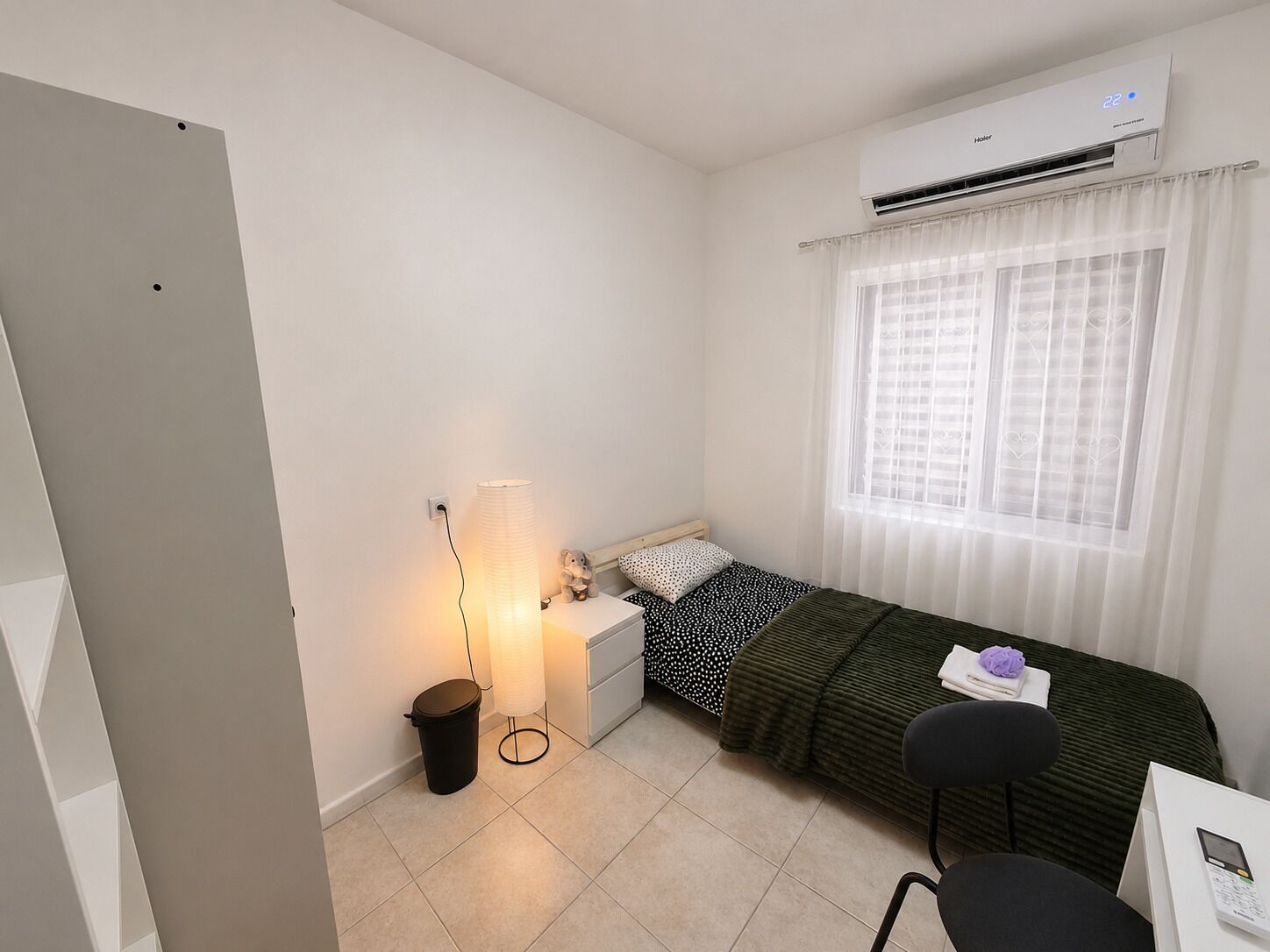A small, tidy room with a bed, a desk, a chair, a trash bin, a lamp, and an air conditioning unit.