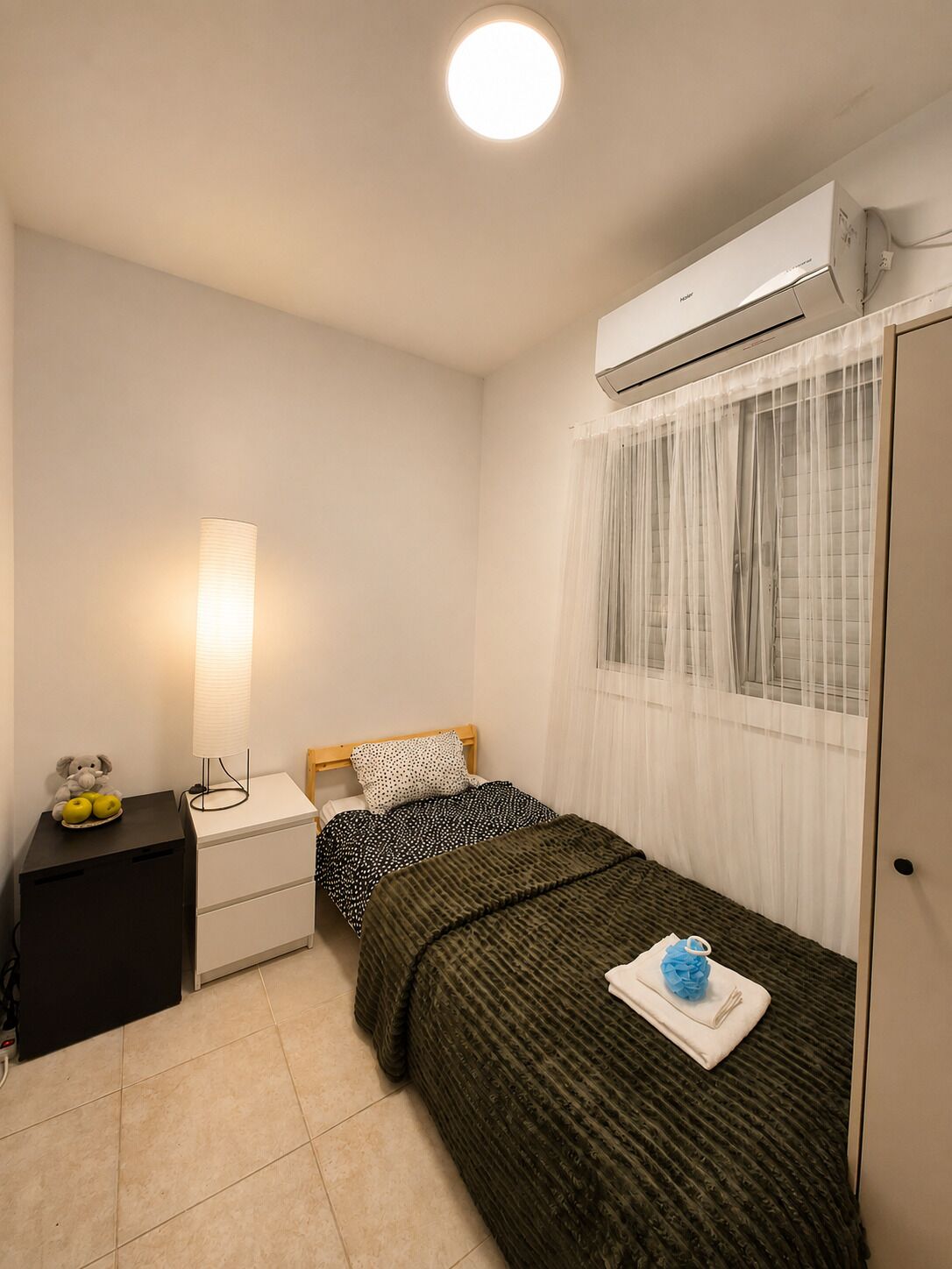 A small, clean room with a bed, a bedside table, a lamp, and an air conditioning unit.