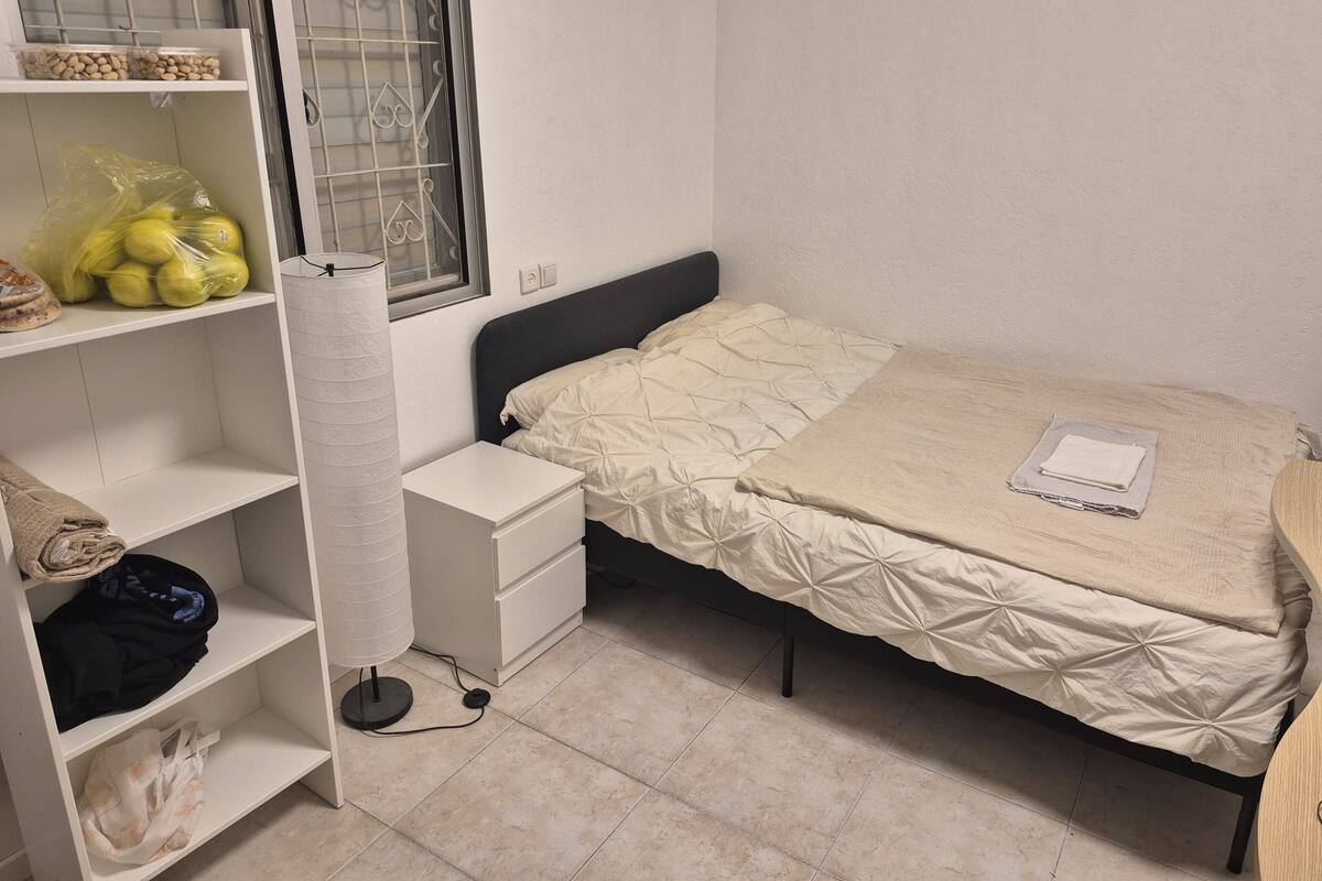 Un dormitorio con una cama, una mesita de noche, una estantería con objetos y una ventana con barrotes.