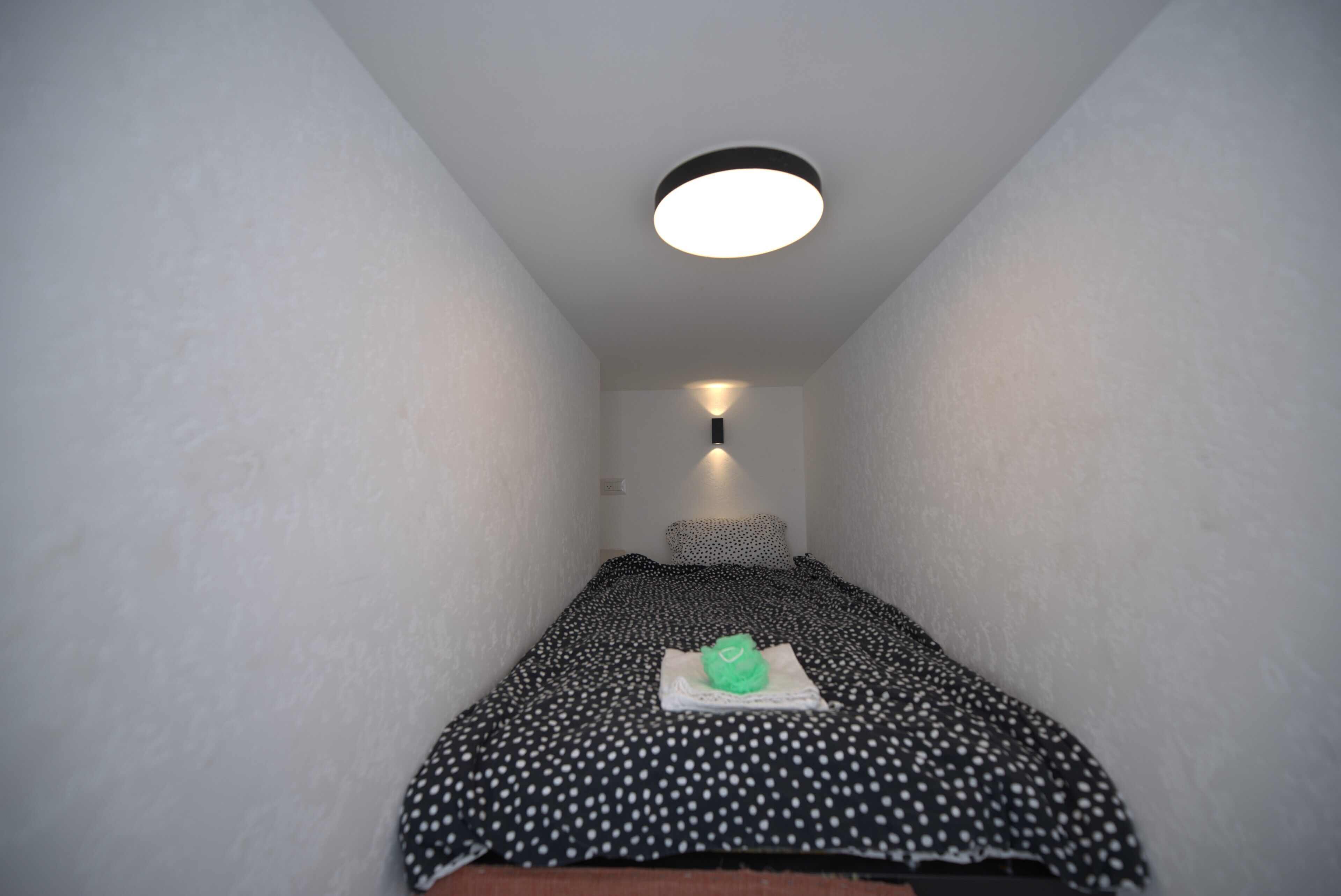 Una habitación estrecha con una cama, una lámpara de pared y una lámpara en el techo.