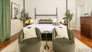A bedroom with a bed, two armchairs, a bedside table, a lamp, a picture on the wall, and a plant.
