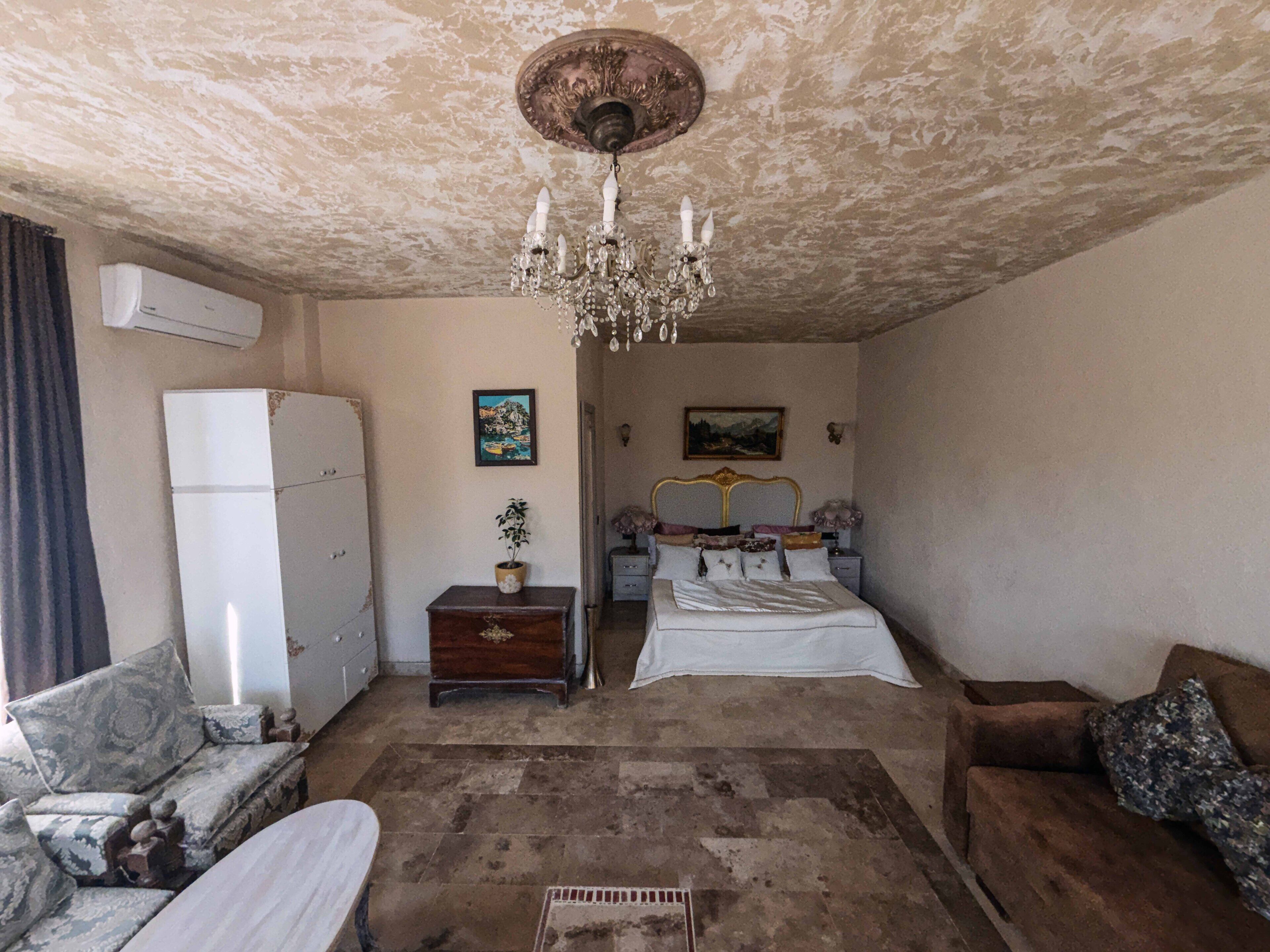 A bedroom with a bed, a dresser, a chest of drawers, a sofa, a small table with a plant, and a chandelier.