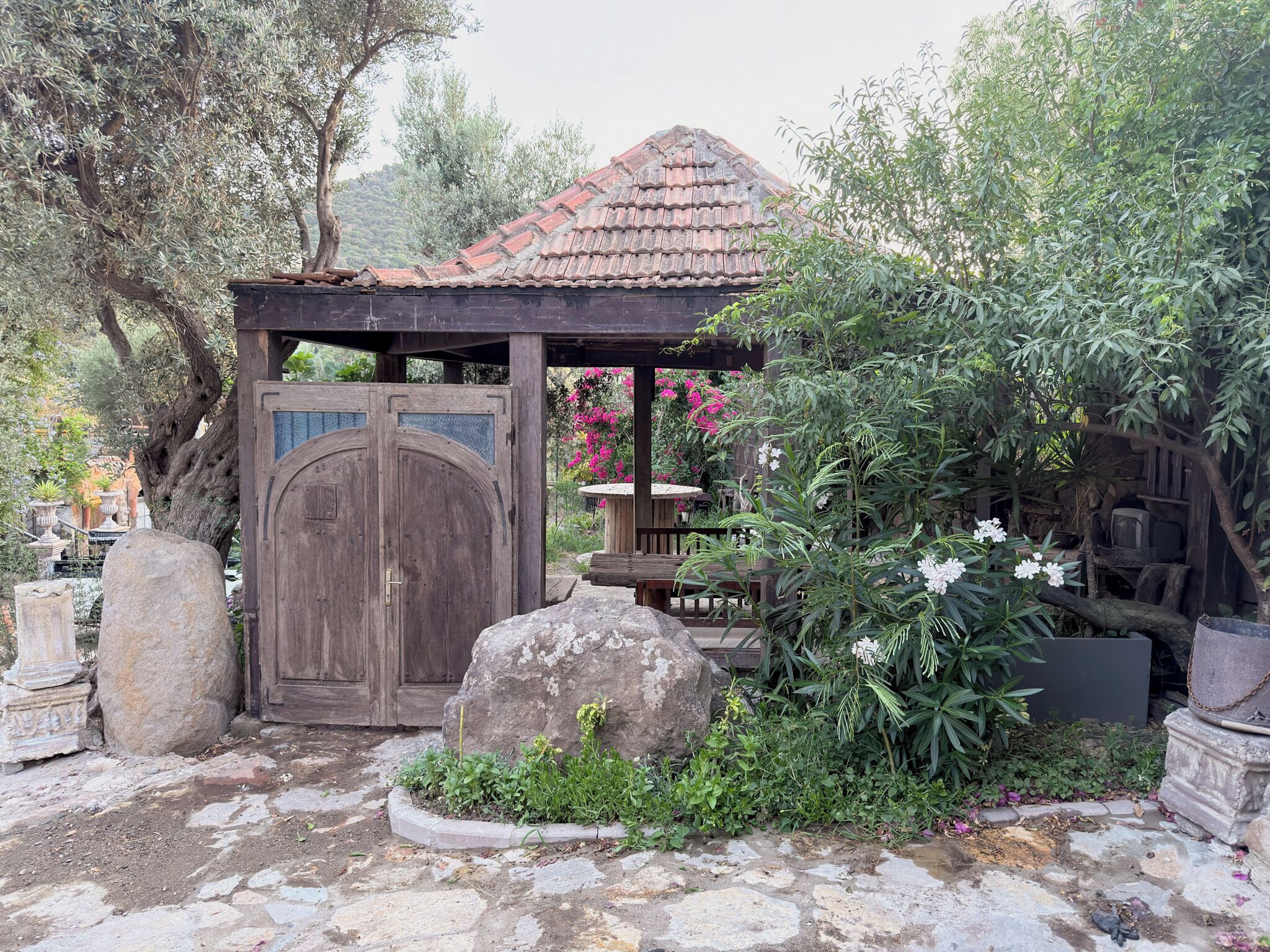 A rustic wooden gazebo with a tiled roof, surrounded by lush greenery and flowering plants.