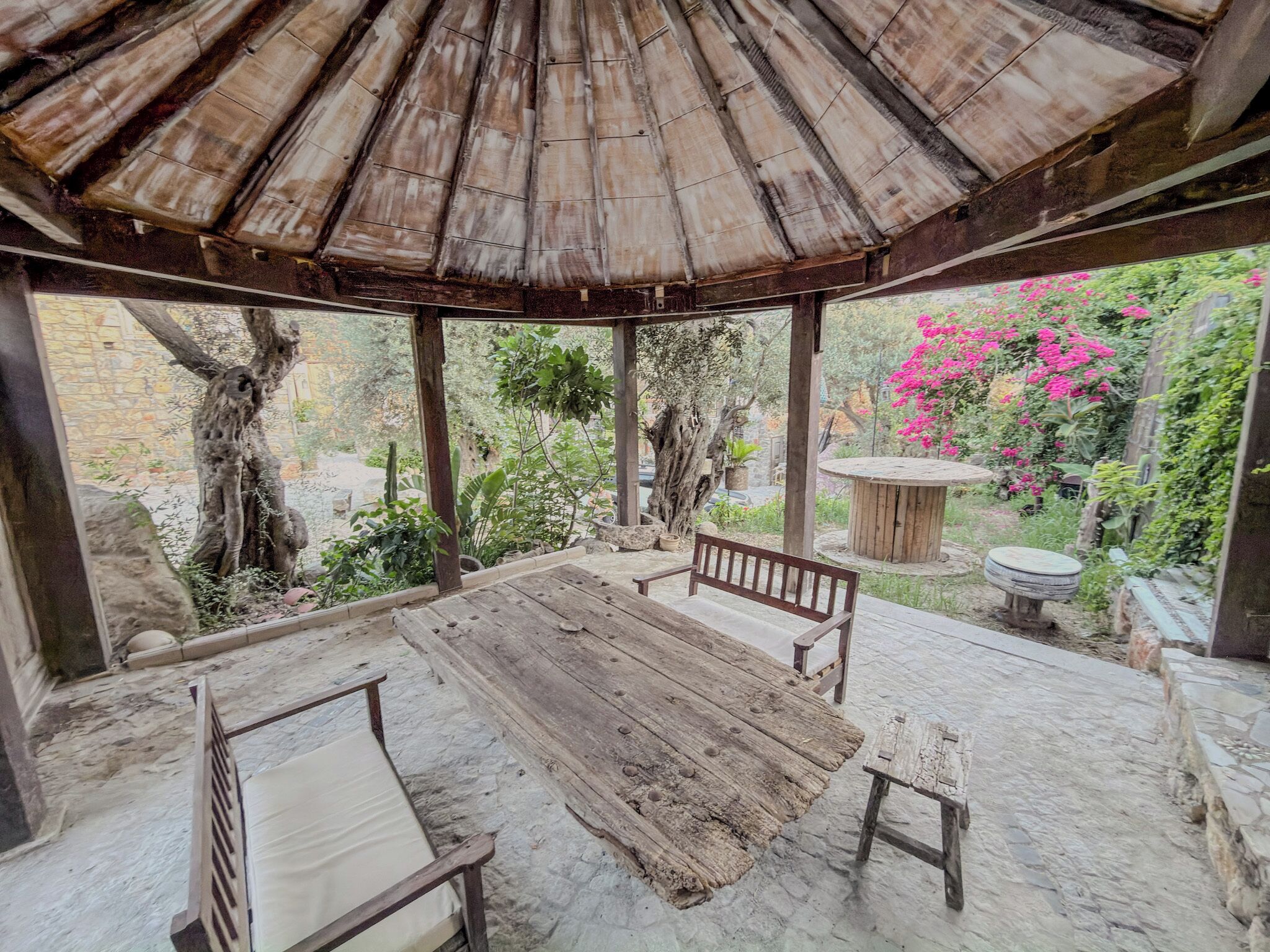A covered outdoor seating area with a wooden table and benches, surrounded by greenery and a stone wall.