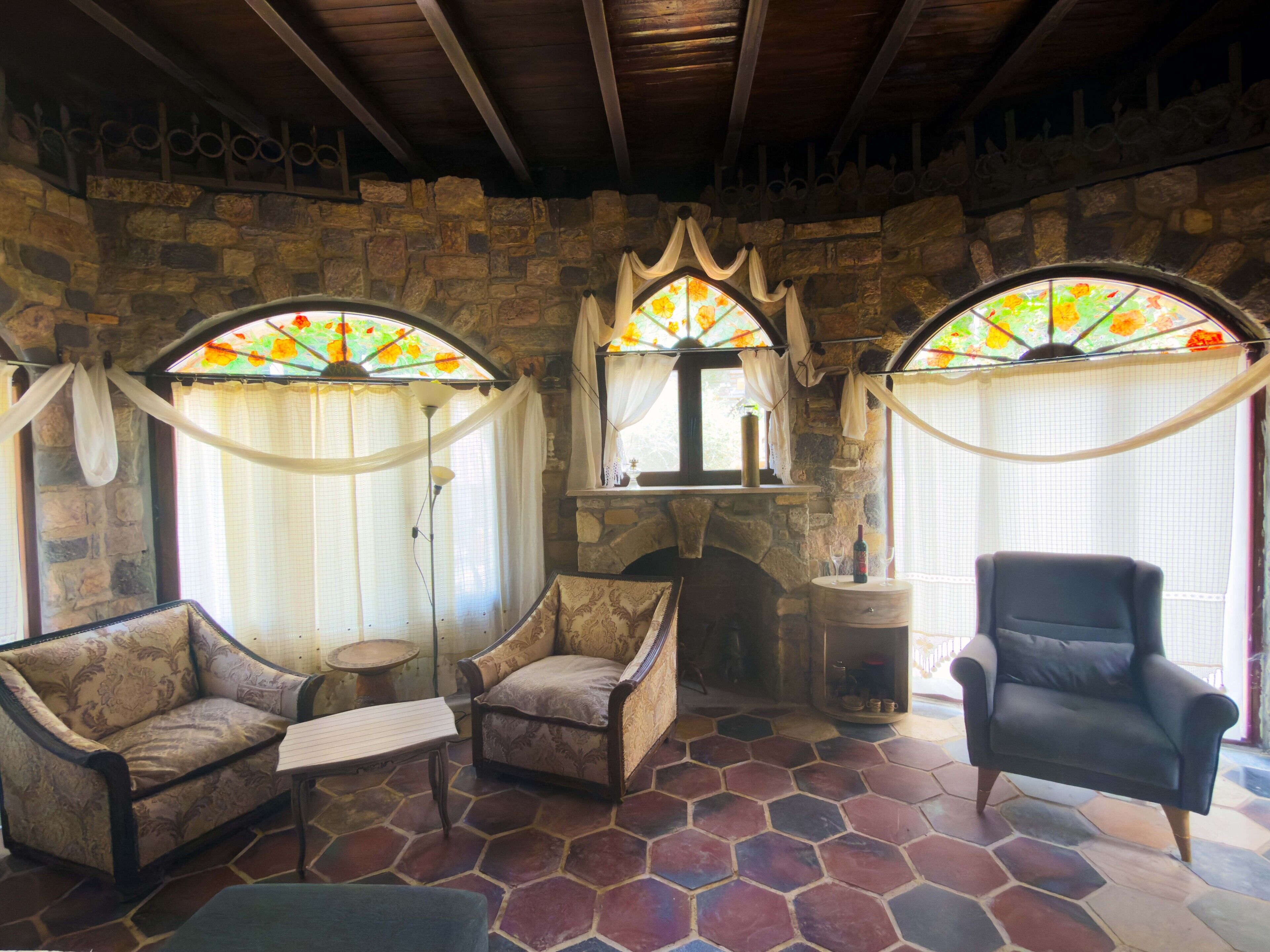 Une pièce aux murs de pierre, dotée d’une cheminée, de deux fauteuils, d’un canapé et d’une grande fenêtre ornée de vitraux.