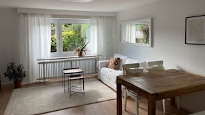 Ein modernes Wohnzimmer mit einem weißen Sofa, einem Holzesstisch, einer Heizung und einem Fenster mit Blick ins Grüne.