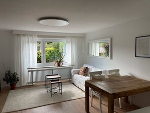 Ein modernes Wohnzimmer mit einem weißen Sofa, einem Holzesstisch, einer Heizung und einem Fenster mit Blick ins Grüne.